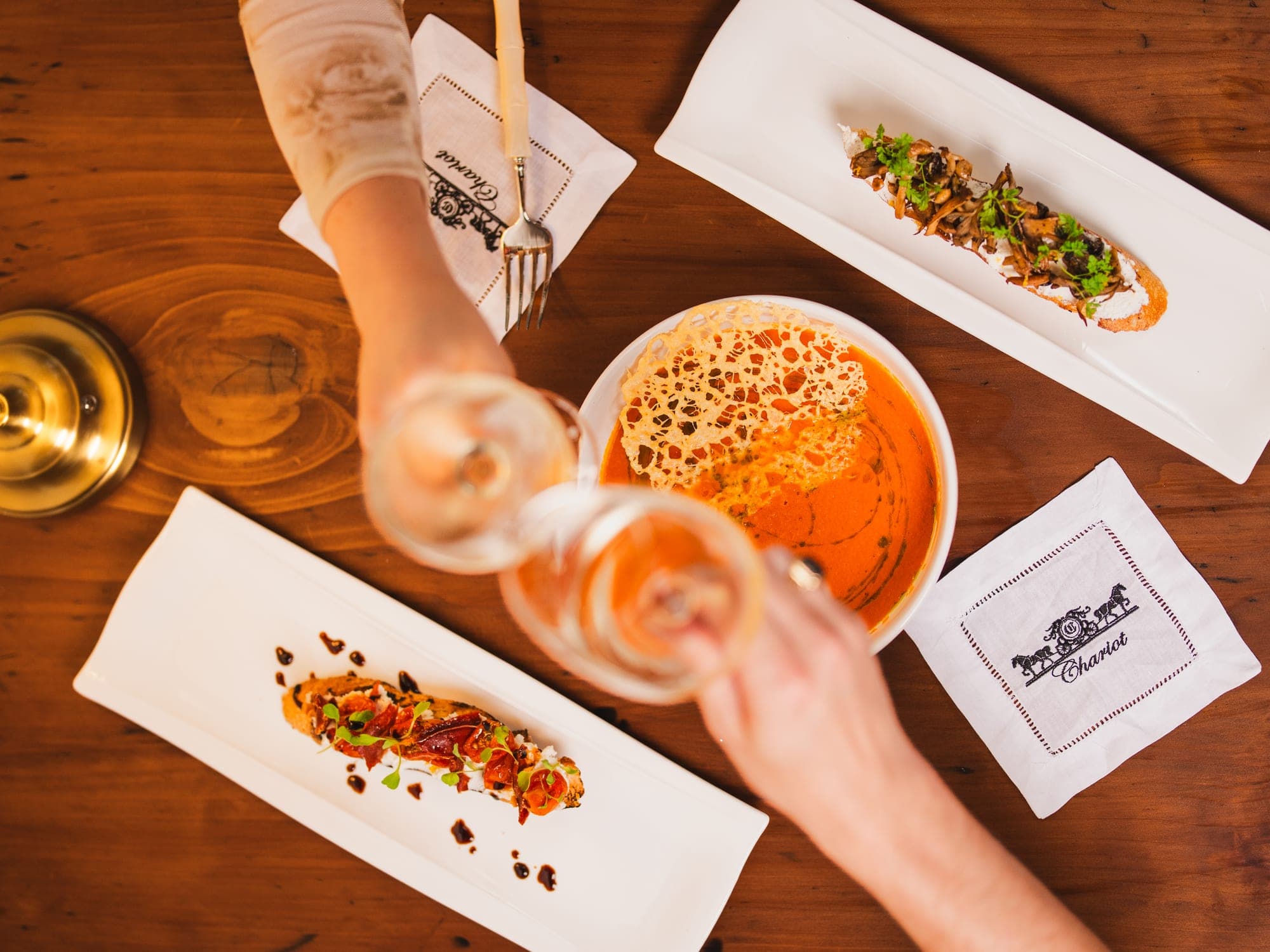 Two glasses meeting over the table at Chariot, toasts and tomato soup laid out between them.